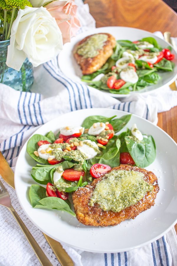 Crispy Chicken Caprese Salad is made with fresh baby spinach topped with sliced tomatoes, buffalo mozzarella, crispy breaded chicken, toasted pine nuts, and a pesto dressing.